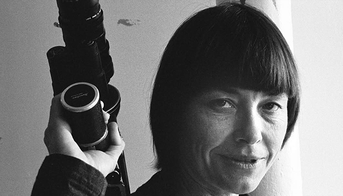 A woman with short hair and bangs holds a camera beside her head.