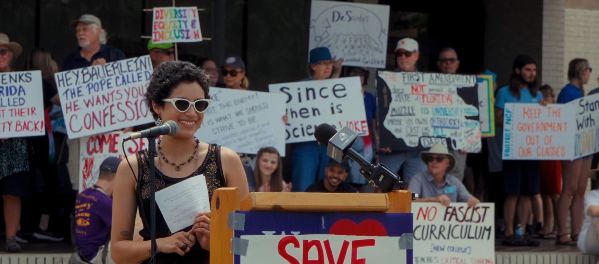 A woman stands at a podium, behind her people stand holding signs.