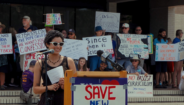 A woman stands at a podium, behind her people stand holding signs.