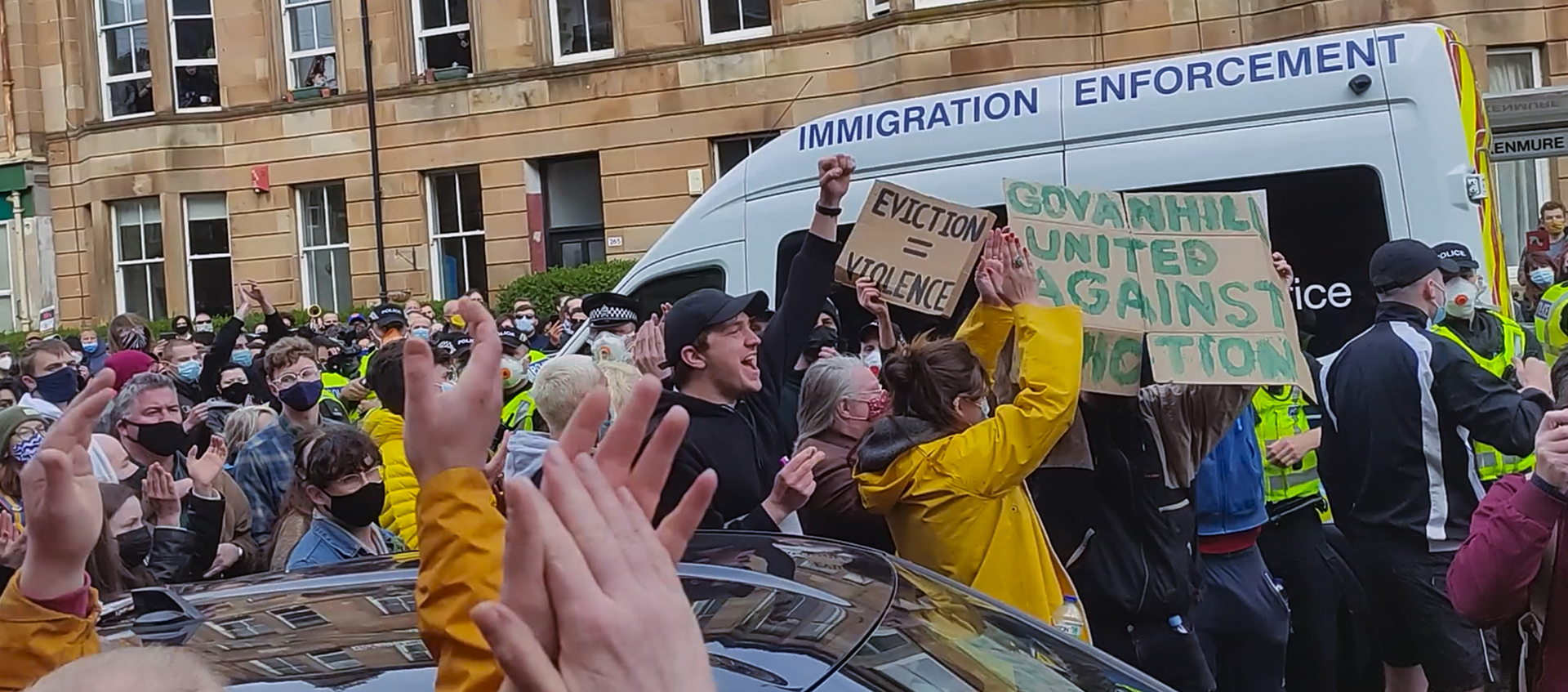 A crowd of people surround an Immigration Enforcement van. They hold signs and have hands and fists in the air.