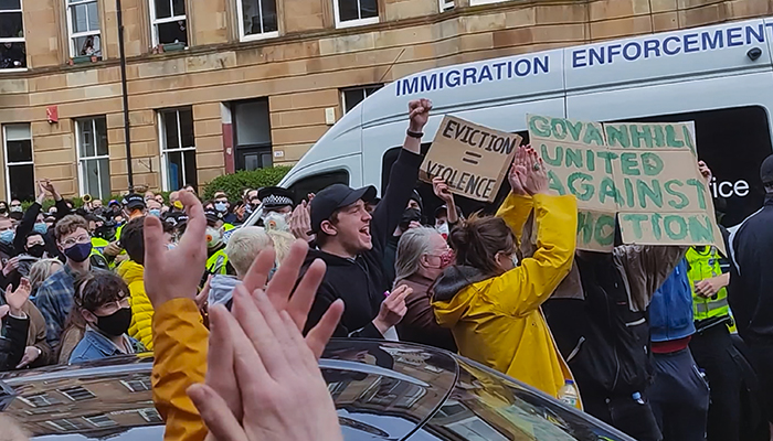 A crowd of people surround an Immigration Enforcement van. They hold signs and have hands and fists in the air.