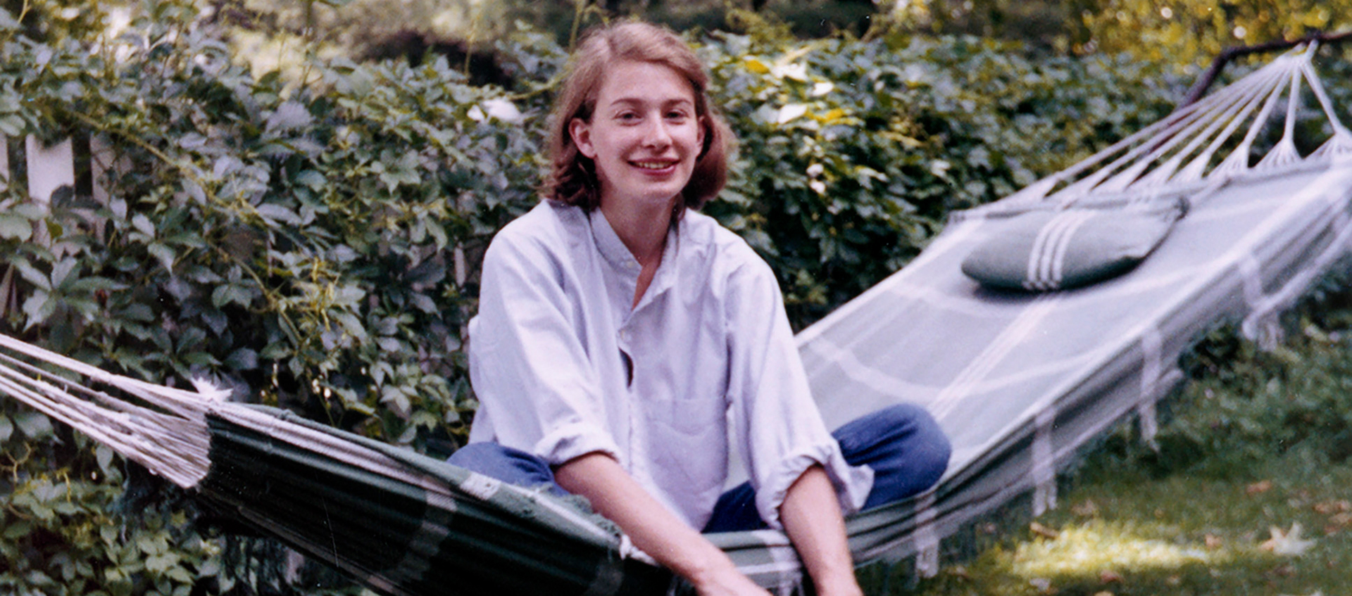 A young woman sits sideways in a hammock in front of a fence covered in greenery.