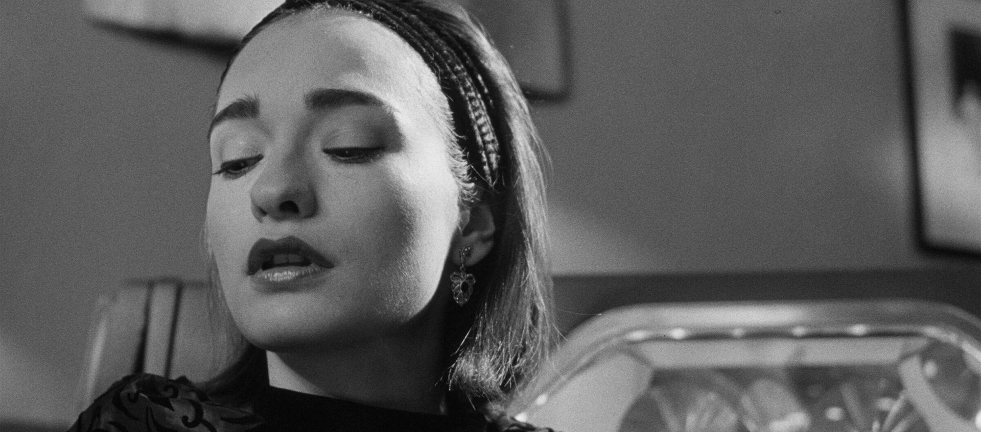 A woman wearing a headband and makeup looks down a jukebox can be seen behind her.