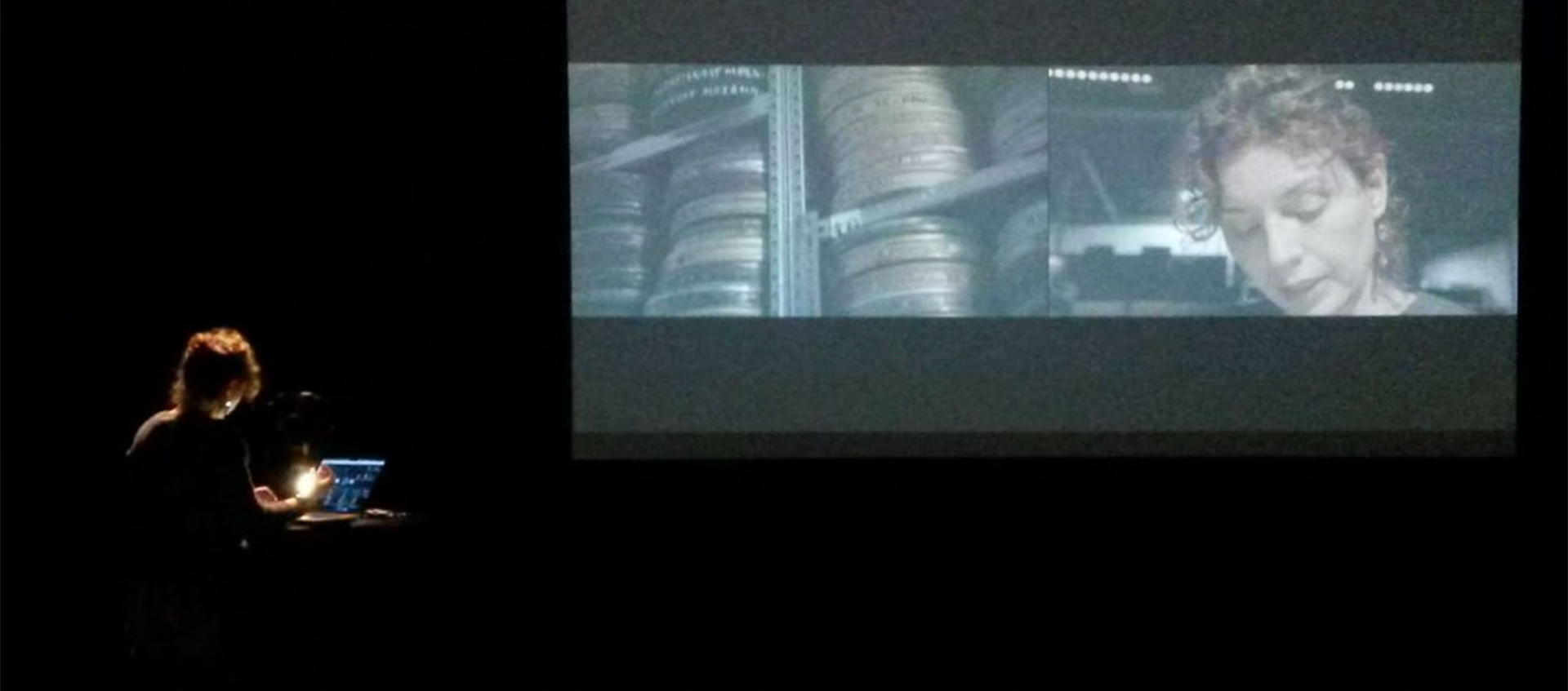 A person stands at a podium with a laptop in a dark room, watching a large screen displaying two film scenes: film reels and a woman's face.