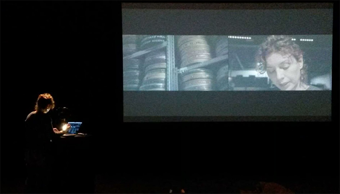 A person stands at a podium with a laptop in a dark room, watching a large screen displaying two film scenes: film reels and a woman's face.