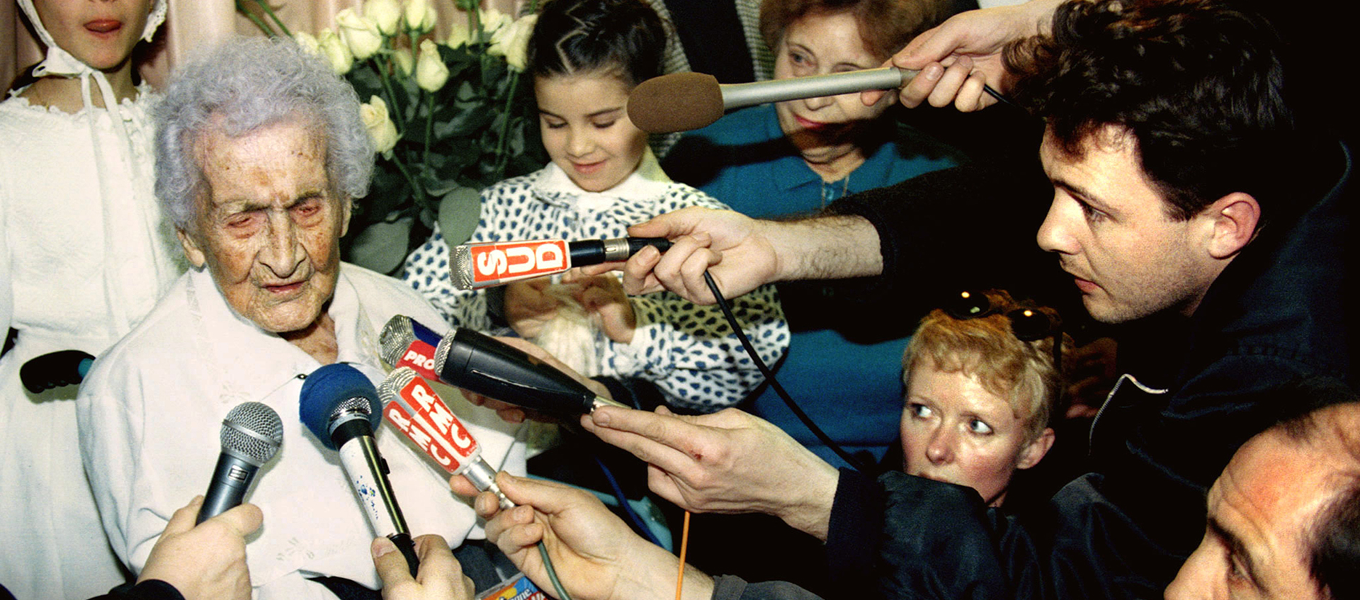 A crowd of people hold microphones from different news agencies up to an elderly woman.