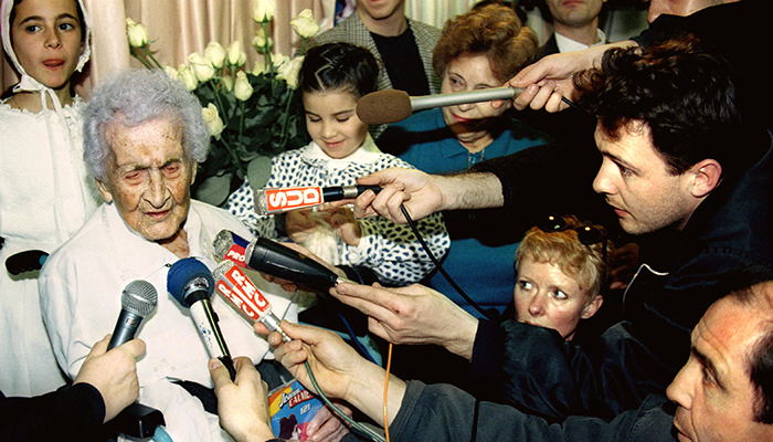  A crowd of people hold microphones from different news agencies up to an elderly woman.