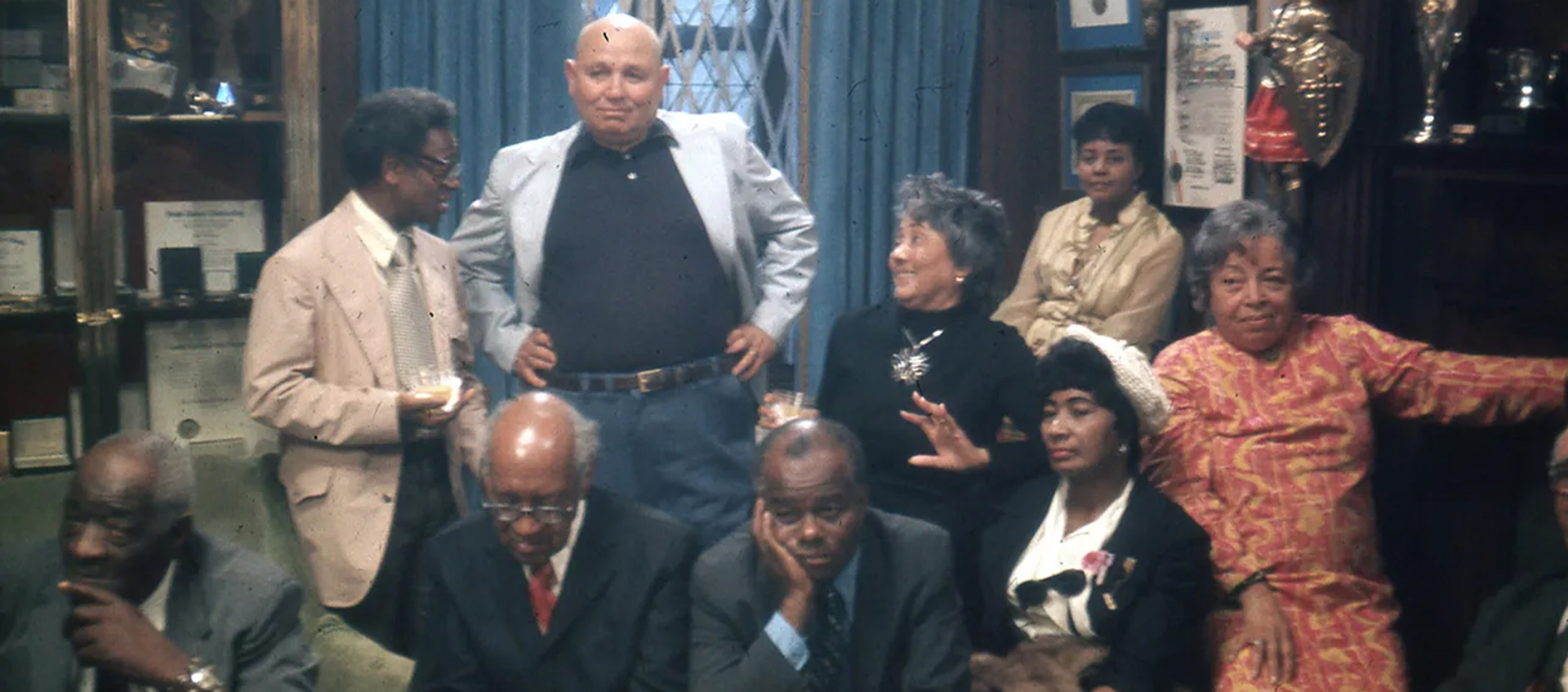A group of nine adults, dressed in formal and semi-formal attire, sit and stand in a wood-paneled room with awards, trophies, and certificates displayed.