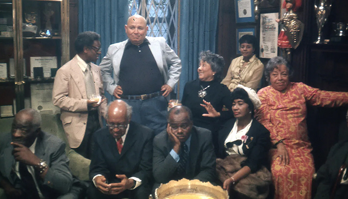 A group of nine adults, dressed in formal and semi-formal attire, sit and stand in a wood-paneled room with awards, trophies, and certificates displayed.
