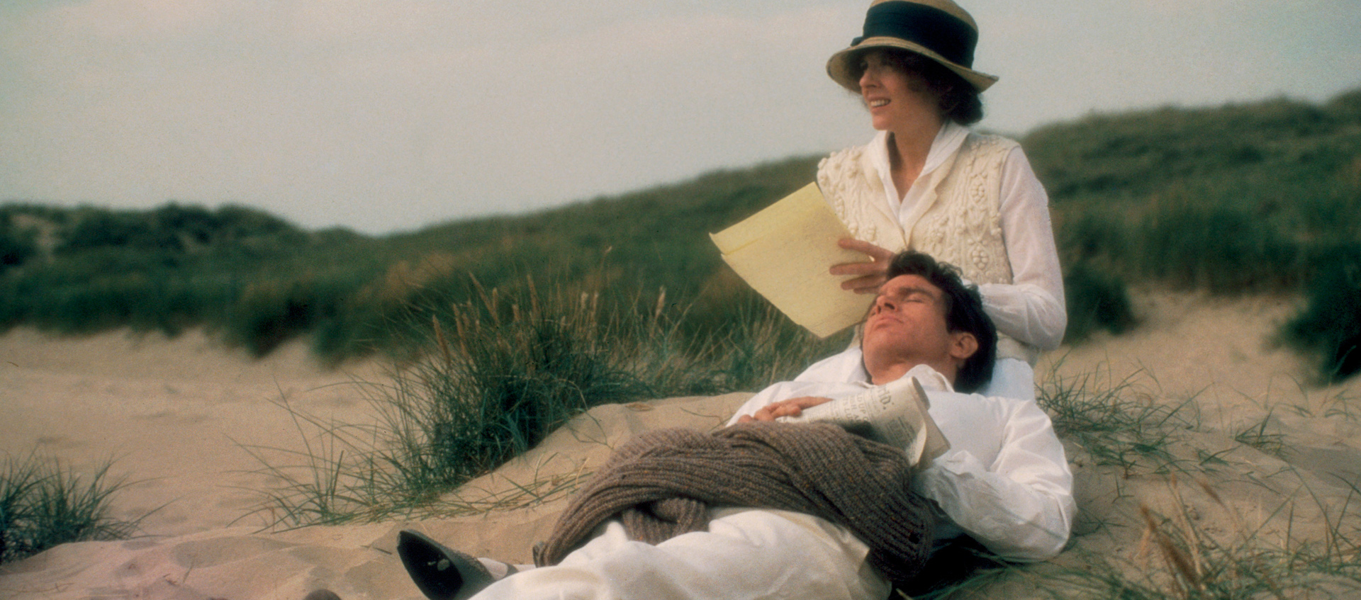 A woman sits while a man lies with his head in her lap. They both wear white clothes and hats, surrounded by grass on a sandy beach.