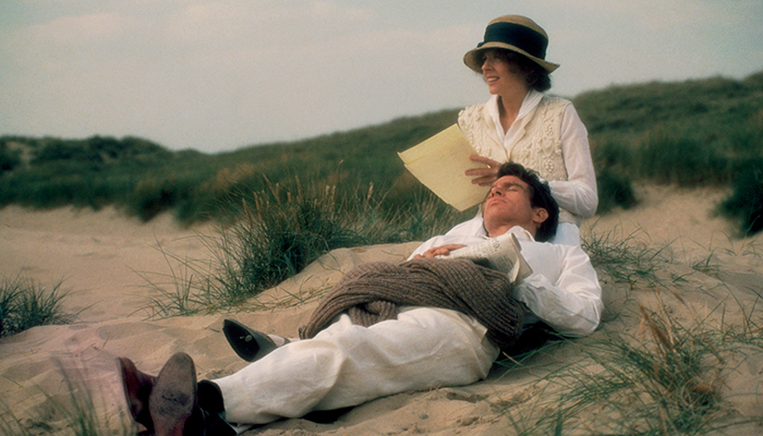 A woman sits while a man lies with his head in her lap. They both wear white clothes and hats, surrounded by grass on a sandy beach.