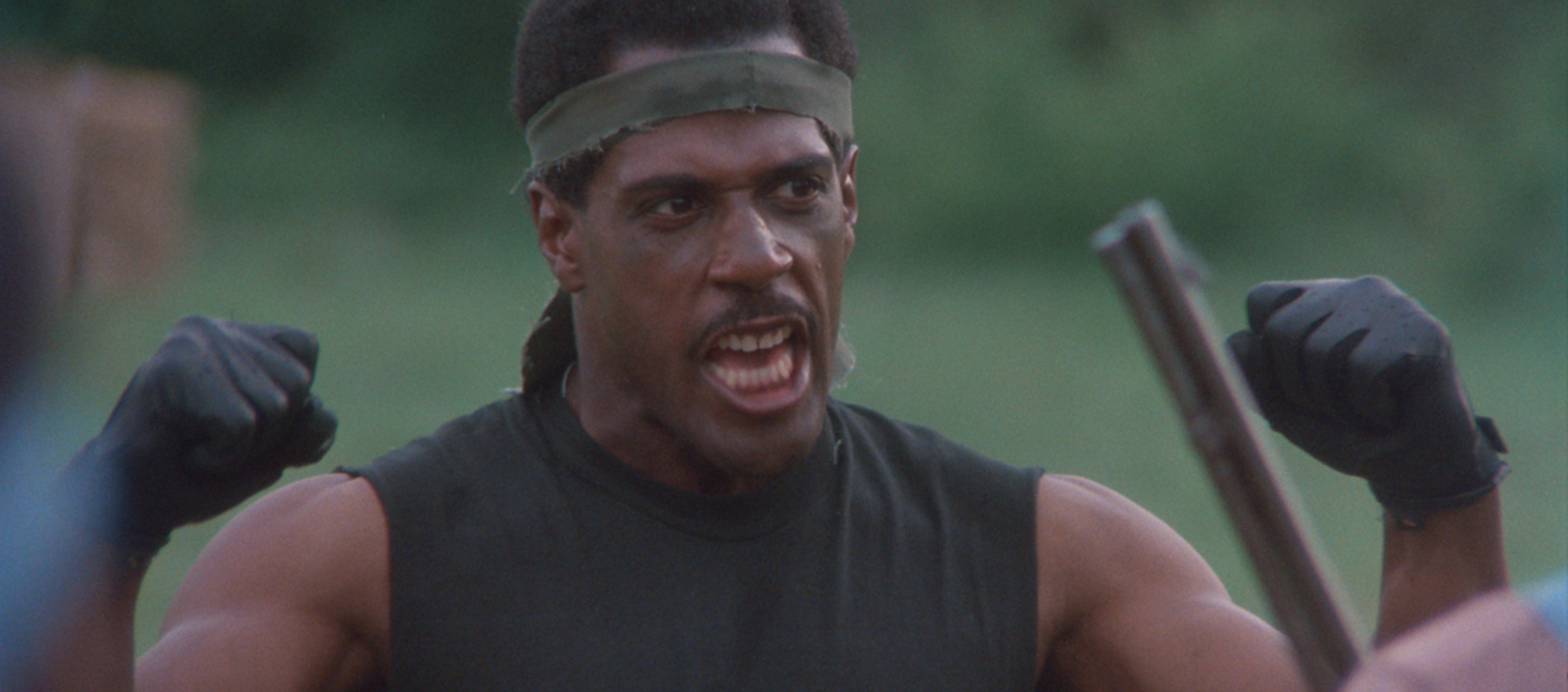 A man pumps his fists in the air. He is wearing a muscle shirt and a headband.