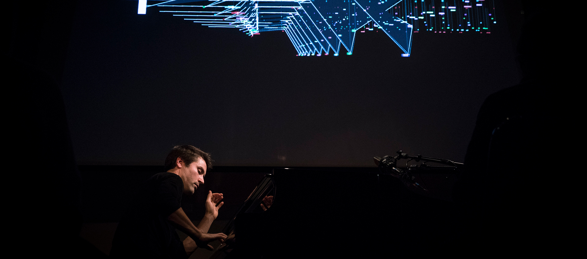 A man plays the piano on a dark stage, above him are blue geometric lights.