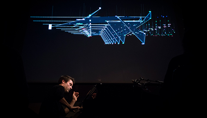 A man plays the piano on a dark stage, above him are blue geometric lights.