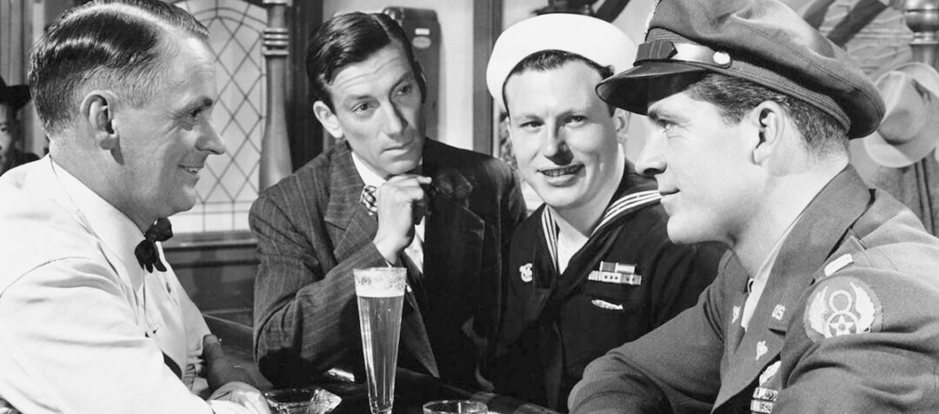 Four men, including a sailor and an airman in uniform, sit at a bar engaged in conversation, with drinks in front of them in a black and white setting.