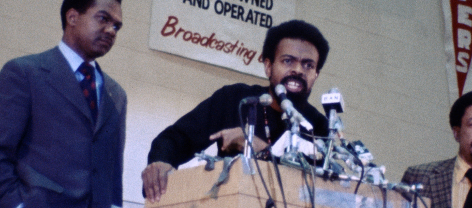 A man speaks into with many microphones at a podium, he is flanked by two other men.