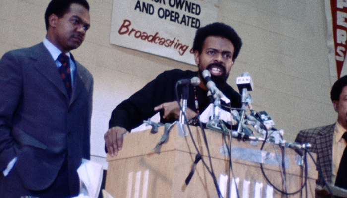 A man speaks into with many microphones at a podium, he is flanked by two other men.