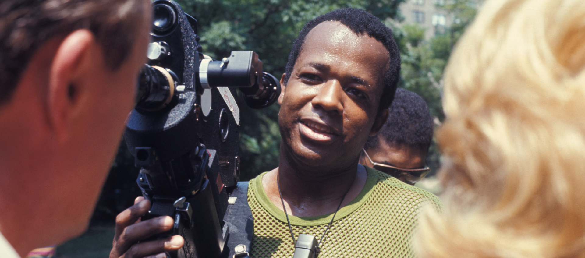 A man holding a film camera stands outdoors, speaking to two people; trees and buildings are visible in the blurred background.