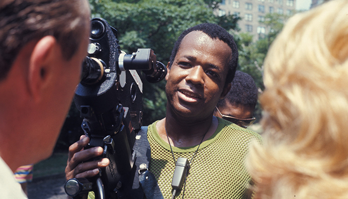 A man holding a film camera stands outdoors, speaking to two people; trees and buildings are visible in the blurred background.