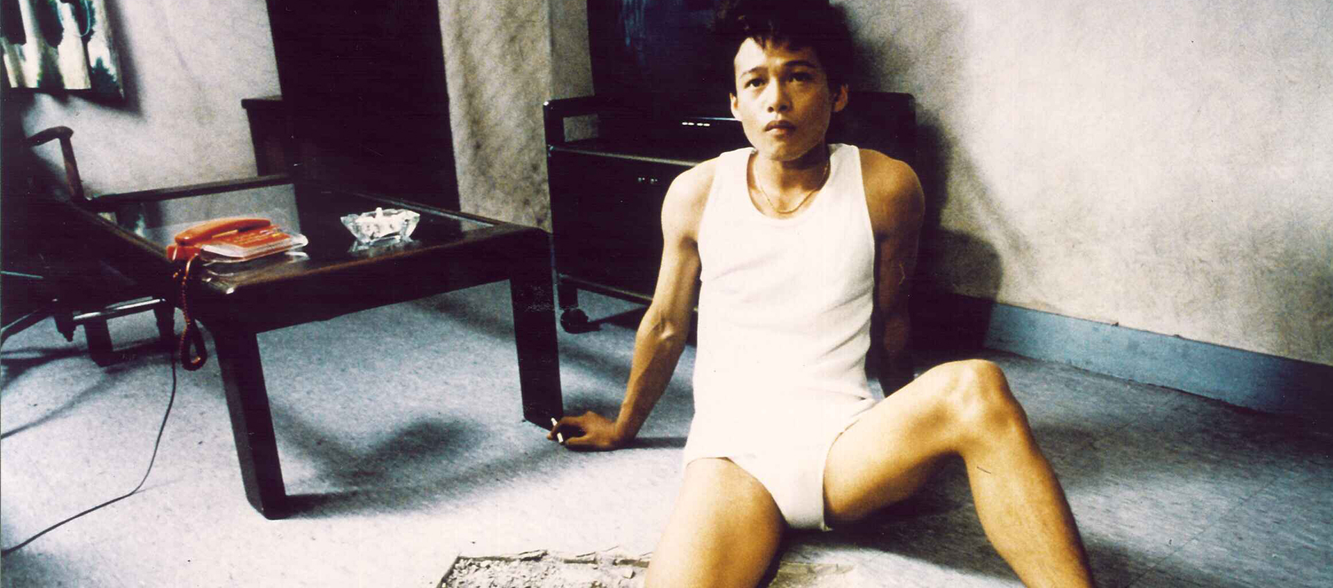 A man sits on the floor in his underwear and undershirt with his leg in a hole in the floor.