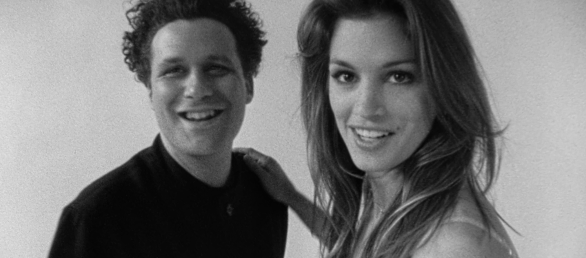 A man with curly hair and a woman with long hair stand close together, both smiling at the camera.