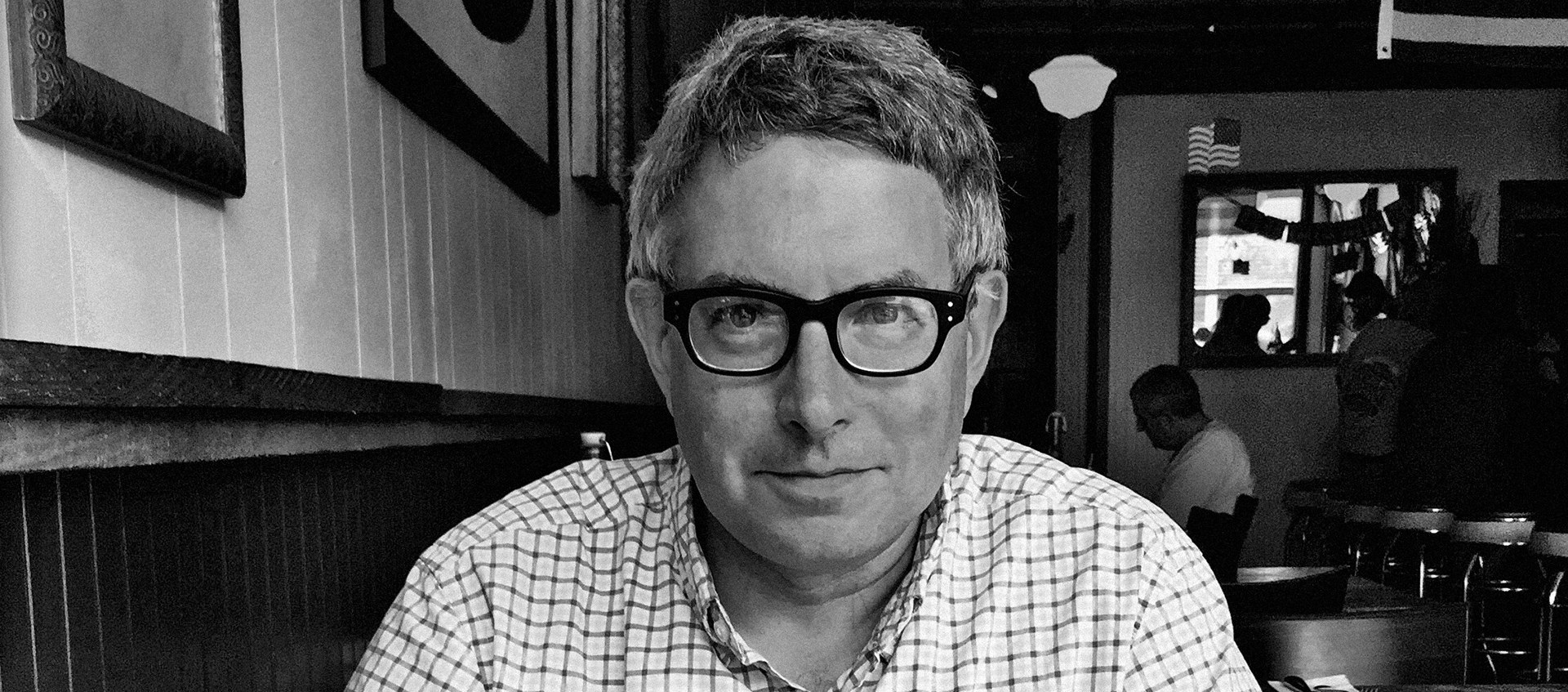 Black-and-white photo of Mark Dion seated in a restaurant. He has gray hair, dark glasses, and wears a plaid shirt. 