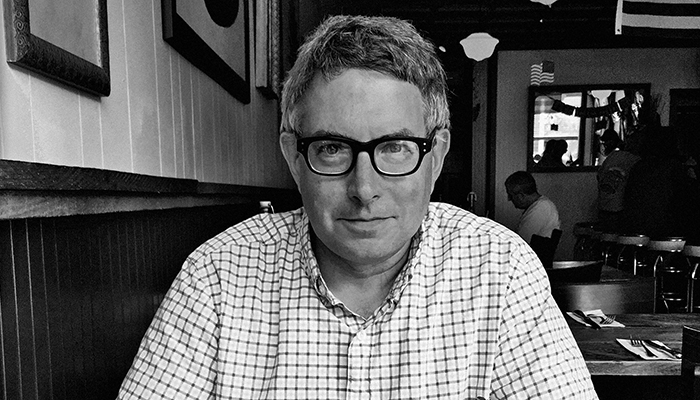 Black-and-white photo of Mark Dion seated in a restaurant. He has gray hair, dark glasses, and wears a plaid shirt. 