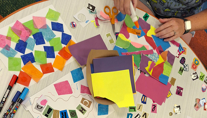 Overhead view of a tabletop featuring collages of colorful paper squares and of irregular torn paper shapes being glued down. 