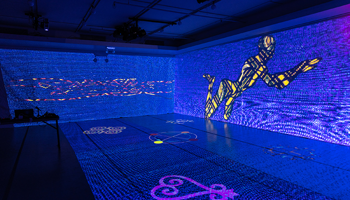 A stage has blue lights and projections of shapes and a person on the floor and the wall.