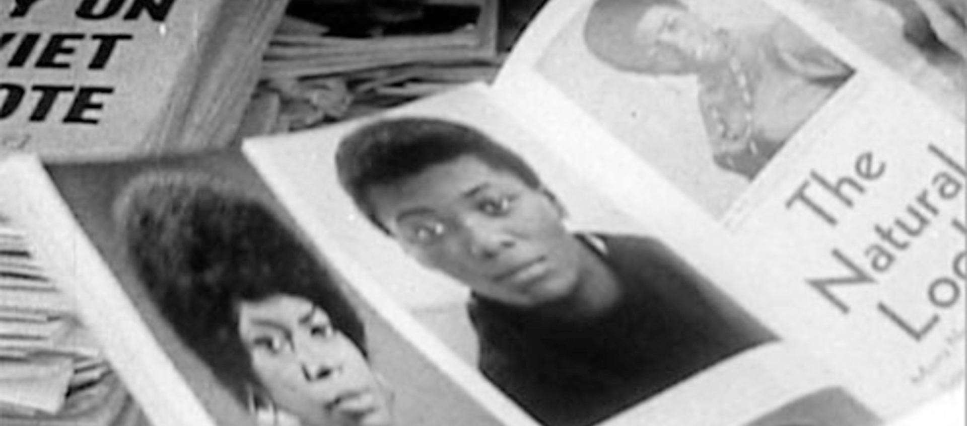 Black and white photo of a magazine open to portraits of Black women with natural hair; papers and a newspaper headline visible in the background.