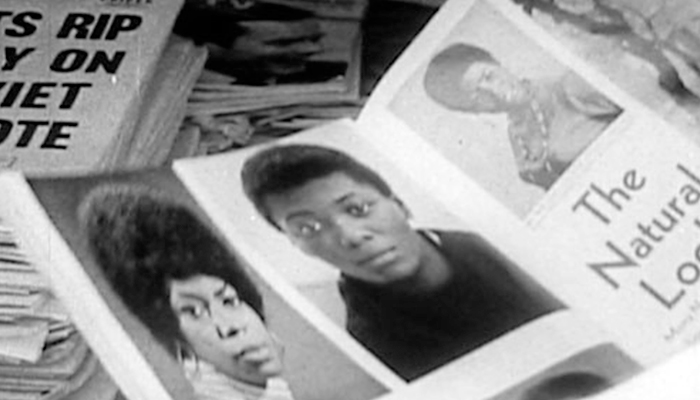 Black and white photo of a magazine open to portraits of Black women with natural hair; papers and a newspaper headline visible in the background.