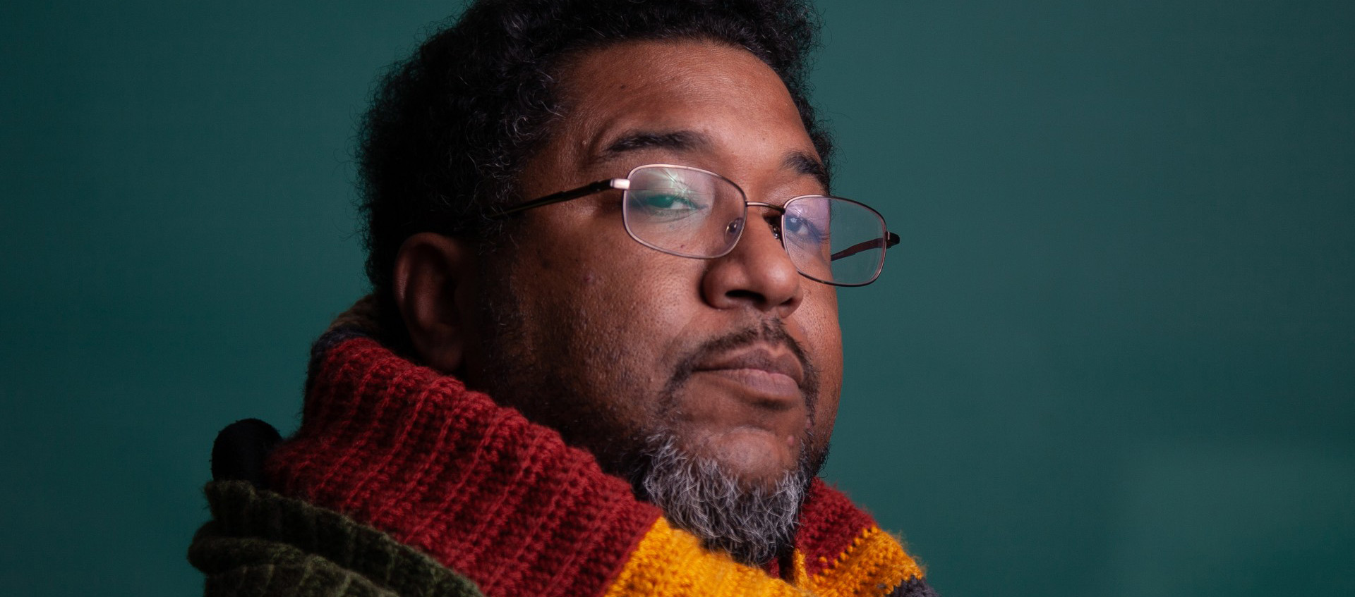 Closeup of a man with medium-brown skin, black hair, and a beard. He is wearing glasses and a multicolored scarf. 
