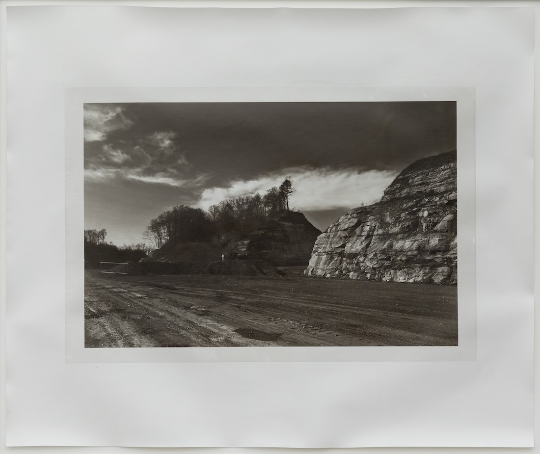 A monochrome image of a landscape with a dirt road with two adjacent rocky structures, one of them covered with trees.
