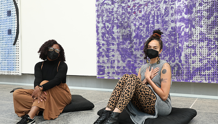 Monique McCrystal (left) and Deja Redman (right) from The Yoga Carriage @ Replenish sit on black cushions on the gallery floor in front of artwork featuring patterns of purple, blue, and white. Monique has black, curly hair, brown skin, glasses, and is wearing a black turtleneck and brown pants. Deja has dark hair in a bun on her head, light brown skin, and is wearing a gray t-shirt and cheetah-print pants. Her hands are crossed on her chest.