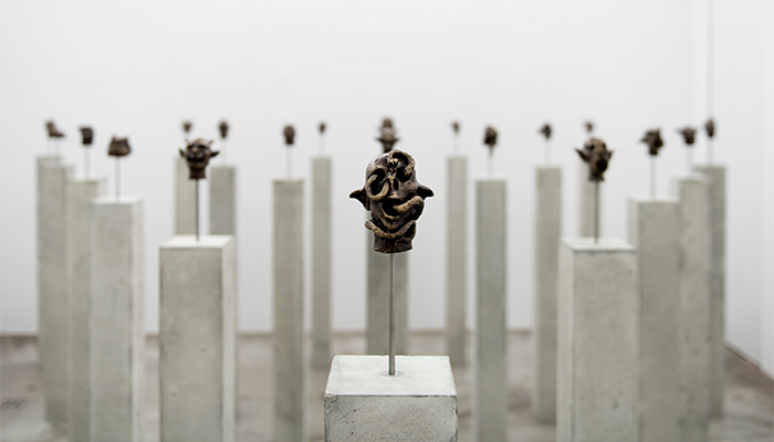 A small bronze sculpture of a head on a steel rod. The goblin-like figure has a snaking passing through its hollow eyes and mouth. Behind it, identical narrow, rectangular concrete pedestals with different bronze heads on each are tightly but evenly spaced in a white gallery.
