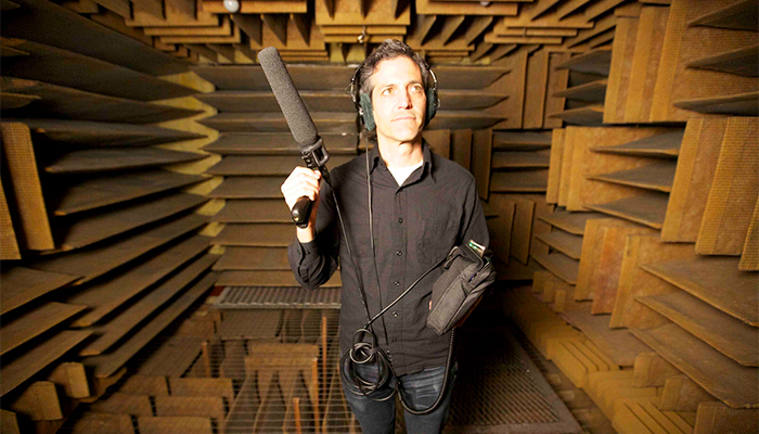 Still of Sam Green, who has light beige skin and dark gray hair; is wearing a black button-down shirt, jeans, and headphones on his ears; and is holding a large microphone with cords hanging from it. He is looking up to the right of the camera and is in an anechoic chamber filled with brown, sound-proof panels that alternate their orientation from vertical to horizontal.