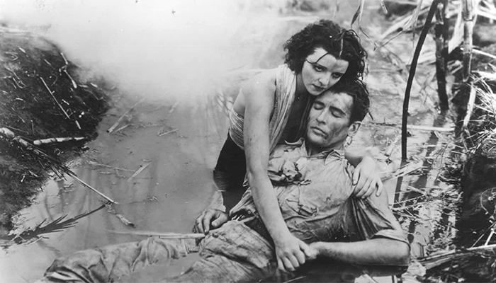 Black-and-white still of a couple with white skin and dark hair. The man wearing jeans and a button-down work shirt is lying unconscious in the debris-filled water, while the woman with a wrap around her neck and bare arms cradles his neck and back and holds his hand. She looks somber and is resting her head against the man’s head.