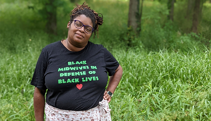 Photo of an adult with brown skin; brown, highlighted, braided hair pulled back in buns; and a black t-shirt with the words “BLACK MIDWIVES IN DEFENSE OF BLACK LIVES” in green text and a red heart underneath. They have one hand on their hip and are smiling slightly, and they are surrounded by tall grass. Tree trunks are in the background.
