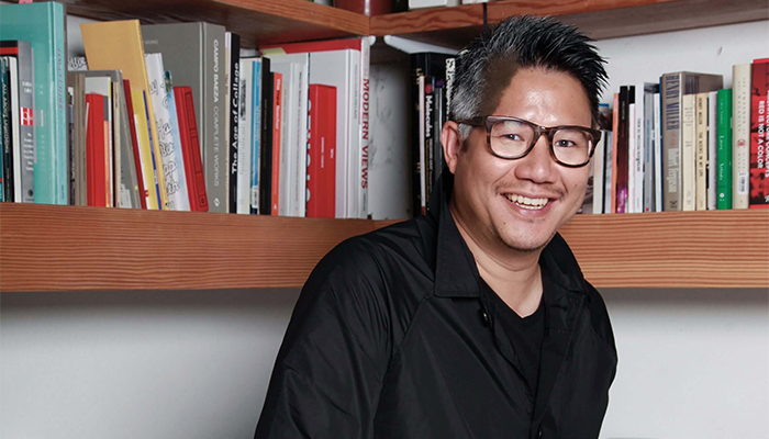 Photo of Kulapat Yantrasast, who has short, black hair with gray on the sides, beige skin, glasses, and a black button-down shirt. He is smiling and standing in front of book-filled, brown wooden shelves protruding from the white wall behind him.