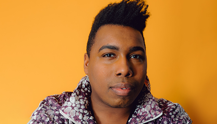 Photo of author Saeed Jones, who has brown skin, short black hair, and a patterned, purple-and-white jacket. They are in front of an orange background.