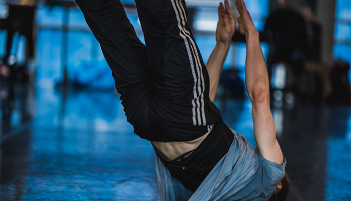 A dancer on their back, with white skin and wearing black pants with white stripes and a blue shirt, extends both arms and legs towards the ceiling. The background is blurred, and blue light is shining through the windows.