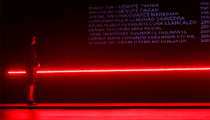 Photo of a person standing to the left but facing the right, holding their hands together in front of them; behind them is a long, hanging beam of red light against a black screen with purple words.