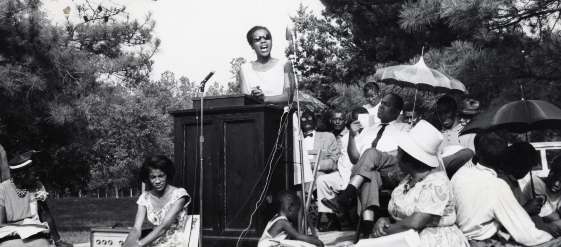Archive black and white photo of activist Ella Baker speaking at a podium in a wooded park, surrounded by Black men, women, and children standing, sitting, and listening