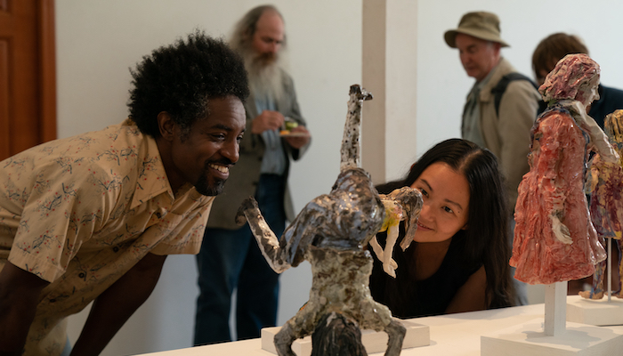 A man and a woman are in a gallery space with other people in the background. The man and woman bend down to look more closely at a ceramic sculpture.